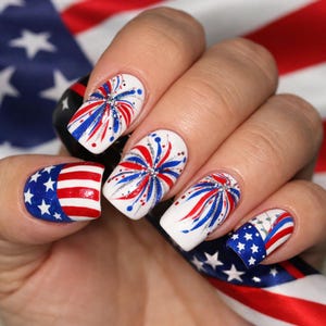 May include: A hand with a manicure featuring patriotic nail art. The nails are painted white with red, white, and blue firework designs and American flag patterns. The background includes an American flag.