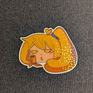 May include: A holographic sticker featuring a stylized cartoon character with yellow hair in a high ponytail, a blue hair tie, and a small yellow heart above the head. The character holds a small orange rectangular device with the text DENZ printed on it.