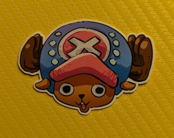 May include: A sticker featuring the character Tony Tony Chopper from the anime series One Piece. The sticker depicts Chopper's face with a pink hat, brown antlers, and a blue hat with a white X symbol.