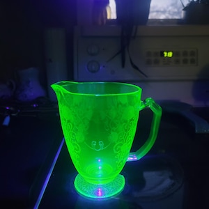 May include: A white ceramic mug with a black handle and the text "But First, Coffee" in a bold, black font. The mug is set against a blurred background, suggesting a cozy setting. The mug appears to be approximately 4 inches tall.