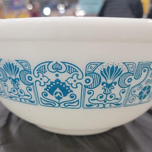 May include: A white milk glass mixing bowl featuring a repeating horizontal band of intricate blue floral and geometric patterns around the exterior. The bowl has a smooth, rounded rim and a tapered base, showcasing a classic mid-century design.