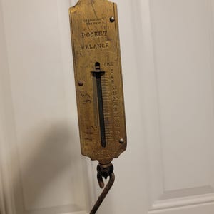 Puede incluir: Una báscula de bolsillo de latón vintage con un anillo de suspensión superior y un gancho inferior. La cara está grabada con el texto FOR SPORTSMEN USE ONLY, POCKET BALANCE, LBS y REGISTERED. Una escala vertical muestra incrementos de 0 a 50.
