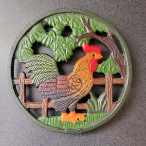 May include: A round cast iron trivet featuring a colourful rooster standing in front of a brown fence. The design includes green foliage, a brown tree branch, and a red comb on the rooster. The rooster has orange, brown, and grey feathers.
