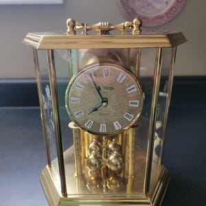 May include: A gold-tone quartz mantel clock with a hexagonal glass case, featuring a gold face with Roman numerals, ornate floral engravings, and a pendulum with four small gold spheres suspended beneath the clock dial, topped with a handle.