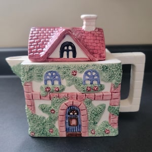 May include: A ceramic teapot shaped like a two-storey cottage with a removable roof lid. The house features white walls, pink brick trim, green ivy vines, blue arched windows, and a blue front door. A white chimney sits atop the pink shingled roof.