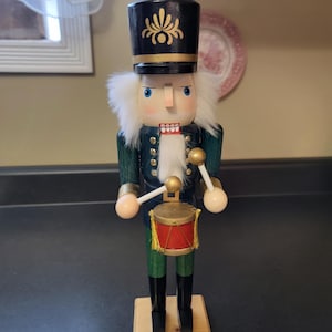 May include: A wooden nutcracker figure wearing a black hat with a gold emblem, a dark green jacket with gold buttons, and black boots. The figure has white faux fur hair and a beard, and is holding two gold-tipped drumsticks over a red drum.