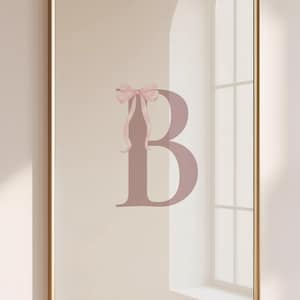 May include: Framed wall art featuring a large letter "B" in a dusty rose color, adorned with a delicate pink bow. The artwork is set against a neutral background and framed in a gold tone. The piece is displayed on a light-colored wall.