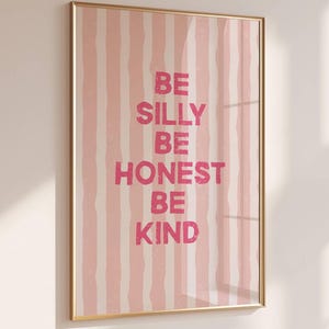 May include: A framed print with a gold frame. The artwork displays a pink and white striped pattern. The words "BE SILLY BE HONEST BE KIND" are written in pink. The print is mounted on a white wall.