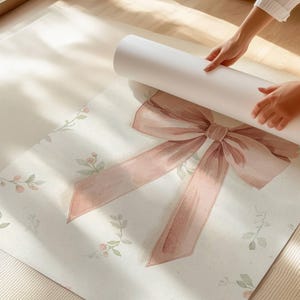 May include: A rolled-up white paper product is held over a large, flat, off-white surface. The surface is decorated with a large, watercolour-style pink bow and delicate floral sprigs. The bow is the focal point, with its soft, shaded details.