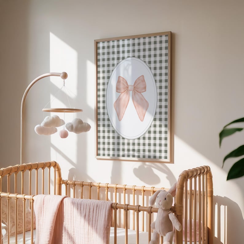 May include: A framed print with a watercolour bow in pale pink, set against a white oval and a grey and white gingham background. The artwork is displayed in a nursery setting, with a wooden cot and mobile.