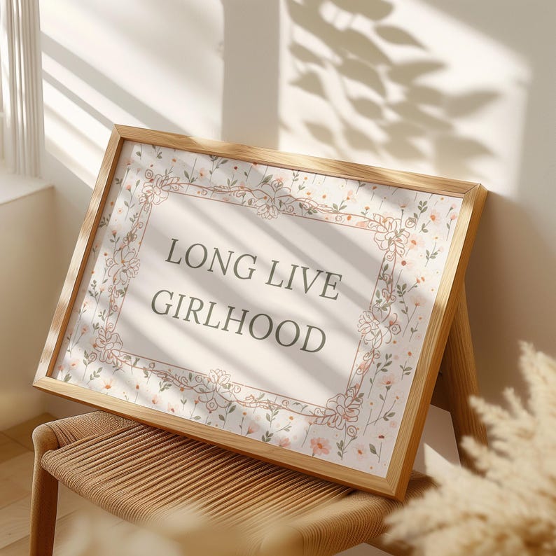 May include: A wooden framed print featuring the words "LONG LIVE GIRLHOOD". The print has a floral design border and a light background. The frame is made of light-coloured wood.