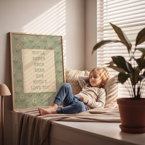 May include: A framed print with the text "YOU'LL NEVER KNOW DEAR HOW MUCH I LOVE YOU" on a floral background. A child is resting on a window seat with pillows and a blanket. A potted plant sits nearby.