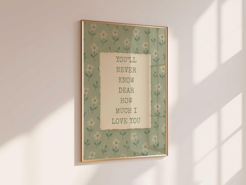 May include: A framed print with a gold-coloured frame. The print features a floral pattern in the background and a central piece of paper with the text "YOU'LL NEVER KNOW DEAR HOW MUCH I LOVE YOU". The background is a light green colour.