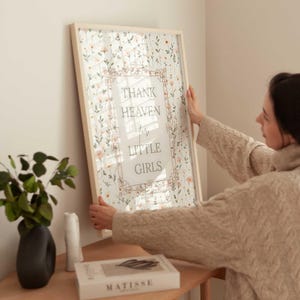 May include: A framed print with the text "THANK HEAVEN for LITTLE GIRLS" in a decorative frame. The print features a floral pattern in soft pinks and greens. The frame is made of light-coloured wood.