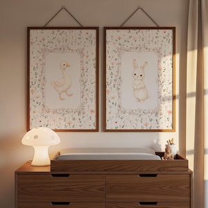 May include: Two framed nursery art prints with a goose and a bunny on a floral background. The prints are in wooden frames, hanging above a wooden chest of drawers with a changing mat. A mushroom-shaped lamp sits on the chest of drawers.