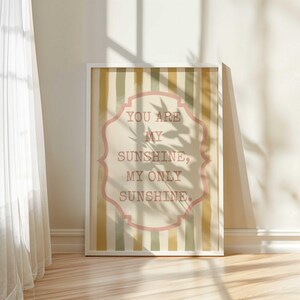 May include: A framed print with the text "YOU ARE MY SUNSHINE, MY ONLY SUNSHINE" in a pink, ornate frame. The background features vertical stripes in shades of beige, green, and yellow. The print is displayed in a white frame.