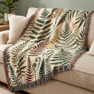 May include: A beige throw blanket with a fern leaf pattern in shades of green, brown, and yellow. The blanket has a fringed edge and is draped over a beige sofa. The botanical design adds a touch of nature to home decor.
