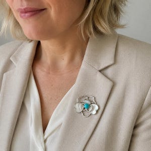 May include: A silver-toned flower brooch featuring white mother-of-pearl petals and a central round turquoise stone. The petals are outlined with a delicate metallic border, creating a layered floral design pinned to a beige blazer.