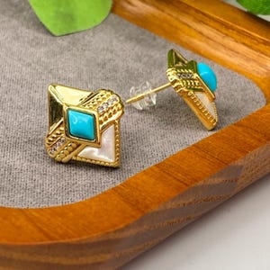 May include: A pair of gold-tone stud earrings featuring a diamond-shaped design. Each earring is accented with a central square turquoise stone, a white triangular inlay, and small clear crystals set along the textured gold-tone metal border.