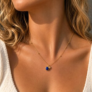 May include: A gold chain necklace featuring small evenly spaced gold beads and a single pendant with a smooth oval blue stone set in a gold frame. The necklace rests against the neck, showcasing the delicate link design and the vibrant blue.