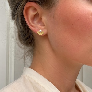 May include: A gold stud earring featuring a small white pearl centered above a textured, semi-circular fan-shaped base. The metal has a matte, granular finish, and the design is symmetrical, with the pearl resting at the top edge of the gold arc.