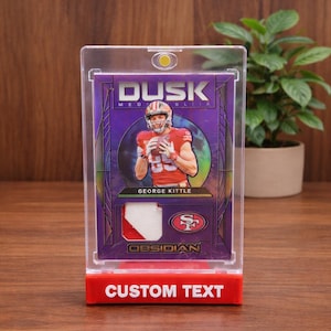 May include: A football trading card of George Kittle in a clear case. The card has a purple and gold design with the word "Dusk" at the top. The bottom of the case has the words "Custom Text" in white on a red base.