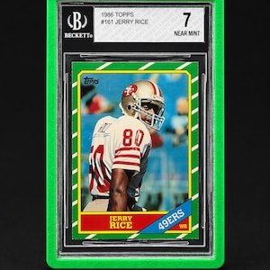 May include: A graded 1986 Topps football card of Jerry Rice, encased in a green protective holder. The card features Rice in his 49ers uniform, with the number 80 visible. The card is graded a 7, labeled "Near Mint".
