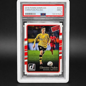 May include: A Panini Donruss trading card featuring Christian Pulisic in a yellow and black Borussia Dortmund uniform. The card is graded MINT 9 by PSA and includes the text "2016 PANINI DONRUSS CHRISTIAN PULISIC". The card is encased in a protective holder.