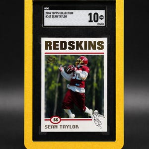 May include: A framed 2004 Topps Collection football card featuring Sean Taylor in a Redskins uniform. The card is graded 10 by SGC. The frame is yellow and the card is in a black holder. The text "REDSKINS" is prominently displayed.
