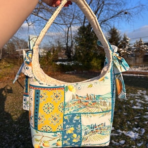 Puede incluir: Bolso de hombro acolchado con un diseño de patchwork colorido. El bolso presenta un asa de color crema y está adornado con paneles de tela que representan limones, paisajes italianos y patrones decorativos. El bolso se sostiene contra un fondo de cielo azul brillante.