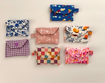 May include: A collection of seven small, fabric coin purses in various patterns and colors. Designs include floral, polka dot, gingham, and geometric patterns. Each purse has a button closure and a small fabric loop.