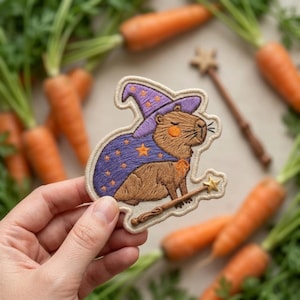 May include: Embroidered patch featuring a capybara dressed as a wizard, wearing a purple hat and cape with gold stars. The capybara holds a wand with a star on the end. Carrots with green tops are in the background.