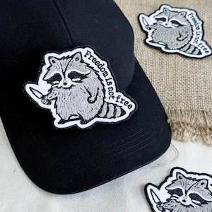 May include: Black baseball cap with an embroidered patch featuring a raccoon holding a knife. The patch has the text "Freedom is not free" in a circular design. Additional patches are visible.
