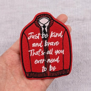 May include: A red embroidered patch shaped like a cardigan sweater with black trim and a white collar. The text "Just be kind and brave That's all you ever need to be" is stitched in white cursive lettering across the front of the garment.
