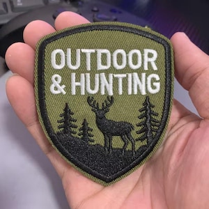 May include: An olive green patch with the words "OUTDOOR & HUNTING" in white. The patch features a black silhouette of a deer between two pine trees. The patch has a black border.