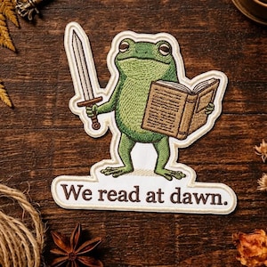 May include: Embroidered patch featuring a green frog holding a sword and a book. The frog is standing on a white banner with the text "We read at dawn." The patch has a detailed design with various shades of green and brown.