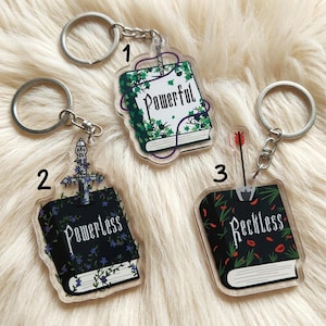 May include: Three acrylic keychains with silver keyrings. Each keychain features a book design with different text: "Powerful", "Powerless", and "Reckless". The "Powerful" keychain has green vines. The "Powerless" keychain has a sword. The "Reckless" keychain has a red arrow.
