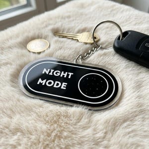 May include: A black and white acrylic keychain with the words "NIGHT MODE" and a starry circle design. The keychain is attached to a silver chain and key ring, with a car key and house key visible.
