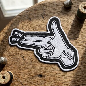 May include: An embroidered patch featuring a white hand forming a finger gun shape against a black background. The text PEW PEW is stitched in white letters on the left side of the patch. The design has a thick black border and white outline.