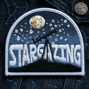 May include: Embroidered patch with a night sky theme. The patch features a crescent moon with gold accents, a telescope, and the word "STARGAZING" in white lettering. The background is a dark blue with white stars.