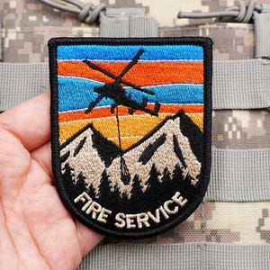 May include: A rectangular embroidered patch with a black border featuring a black helicopter silhouette flying over tan mountains and a forest against a sunset sky of orange and blue stripes. The text FIRE SERVICE is embroidered in tan at the bottom.
