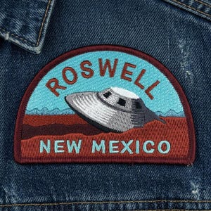 May include: Embroidered patch featuring a gray UFO over a brown landscape, with the text "ROSWELL NEW MEXICO" in teal and red. The patch is sewn onto a blue denim jacket.