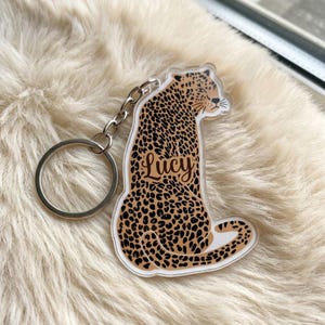 May include: A clear acrylic keychain featuring a leopard illustration with a tan body and black spots. The name Lucy is printed in a dark brown cursive script across the center of the leopard's body. A silver metal split ring is attached to the top.