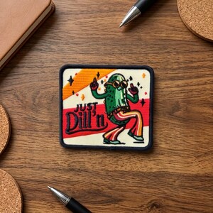 May include: A rectangular embroidered patch featuring a green pickle character wearing sunglasses and striped bell-bottom pants. The design includes the text "Just Dill'n" in dark blue script, set against a cream background with red and orange stripes.