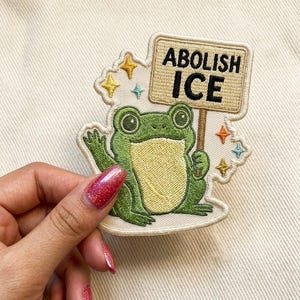 May include: Embroidered patch featuring a green frog holding a sign that reads "ABOLISH ICE." The frog is surrounded by yellow and blue star accents. The patch is on a cream-colored background.