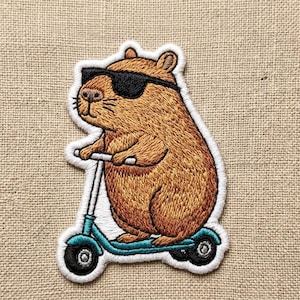 May include: Embroidered patch featuring a brown capybara wearing black sunglasses, riding a teal scooter. The patch has a white border and is set against a neutral fabric background. The capybara is the central focus of the patch.