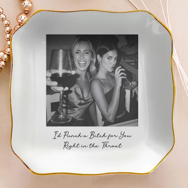 May include: White ceramic trinket dish with a gold rim and a black and white photo of two women. The photo is centered on the dish and has the text "I'd Punch a Bitch for You Right in the Throat" below it. A gold beaded necklace is visible in the upper left corner.