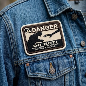 May include: A denim jacket with a rectangular patch featuring a black dinosaur silhouette and the words "DANGER DO NOT! FEED, TEASE, OR APPROACH ANY WILD DINOSAURS." The patch has a brown border and is sewn onto the blue jacket.