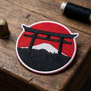 May include: A circular embroidered patch featuring a black torii gate set against a vibrant red background. Below the gate, a black mountain with a white snow-capped peak is depicted. The patch has a white border and is stitched with dense thread.