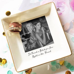 May include: A square, off-white trinket dish with a gold rim, featuring a black and white photo of two women and the text "I'd Punch a Bitch for You Right in the Throat." A rose gold ring sits on the dish.